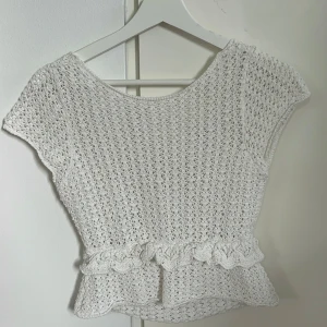 Vit virkad topp med volang och knyt - Superfin vit virkad topp med korta ärmar och volangdetalj i midjan. Toppen har en snygg öppen rygg med knyt baktill. Perfekt till sommaren och passar till både jeans och kjol. Materialet är mjukt och luftigt, perfekt för varma dagar.