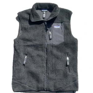 Svart fleeceväst från Patagonia - Mysig svart fleeceväst från Patagonia med hög krage och tre fickor med dragkedja, inklusive en bröstficka. Västen har en klassisk, enkel design och är perfekt att slänga på sig över en hoodie eller tröja när det är kyligt.
