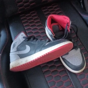 Nike Air Jordan 1 Mid svart/röd/grå - Säljer ett par Nike Air Jordan 1 Mid i svart, grått och rött. Skorna har klassisk Jordan-design med svart snörning, röd insida och röd yttersula. Ovandelen är i skinn och syntet, med perforerad tåbox och ikonisk Swoosh-logga på sidan.