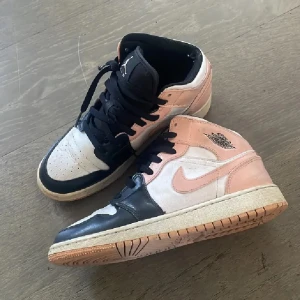 Nike Air Jordan 1 Mid rosa/svart/vit - Nike Air Jordan 1 Mid sneakers i en snygg färgkombo av ljusrosa, svart och vitt. Klassisk siluett med svart snörning, perforerad tå och ikoniska Jordan-loggan på sidan. Ovandel i skinn och syntet, mellanhög modell med bekväm platt sula.