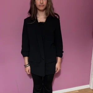 Svart kavaj från Bodyflirt - Stilren svart kavaj från Bodyflirt i klassisk modell med långa ärmar och snygg krage. Kavajen har två fickor framtill och är lätt oversized för en chill look. Perfekt att slänga över en t-shirt eller topp för en clean outfit.