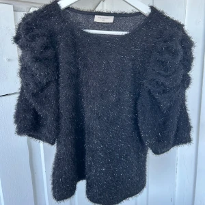 Glittrig svart topp från Freequent - Säljer en svart glittrig topp från Freequent i storlek S. Toppen har korta puffärmar och är gjord i ett fluffigt, syntetiskt material med glansiga trådar som ger en festlig vibe. Perfekt för dig som gillar att sticka ut med coola detaljer och shimmer.