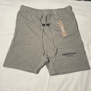  Essentials FOG shorts - Snygga grå shorts från Essentials Fear of God med elastisk midja och snörning. Mjukt bomullsmaterial och diskret logga på benet. Perfekta för chill dagar eller när du vill ha en clean streetwear-look.