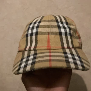 Burberry rutig keps i bomull - Snygg keps från Burberry med klassiskt rutigt mönster i beige, svart, vitt och rött. Justerbar baktill med svart spänne och tydlig Burberry London England-logga. Tillverkad i bomull och perfekt för dig som vill sticka ut med en ikonisk accessoar.