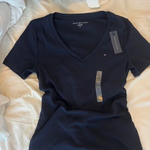 Mörkblå NY v-ringad t-shirt Tommy Hilfiger - Marinblå från Tommy Hilfiger med v-ringning och liten broderad logga på bröstet. Klassisk passform och korta ärmar. Tillverkad i mjuk bomull som känns skön mot huden. Perfekt för en clean och stilren look.