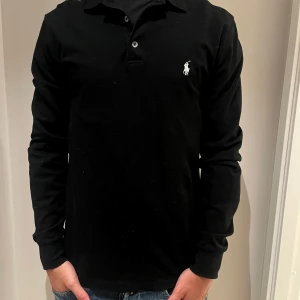 Svart långärmad pikétröja Ralph Lauren - Klassisk svart långärmad pikétröja från Ralph Lauren med vit broderad logga på bröstet. Tröjan har krage, knappar vid halsen och är gjord i mjuk bomull. Perfekt för en clean och stilren look. Aldrig använd ba legat i min garderob. 