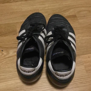 Adidas Copa Mundial fotbollsskor svart/vit - Klassiska Adidas Copa Mundial fotbollsskor i svart skinn med vita detaljer och de ikoniska tre ränderna på sidorna. Skorna har snörning och en platt sula, perfekt för dig som vill ha retrovibbar på planen. Säljs för att de är förr små på mig nu. Priset kan sänkas.