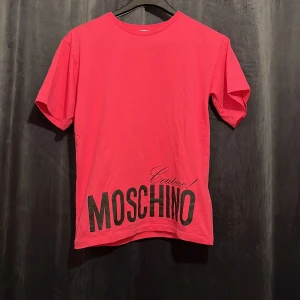 Rosa Moschino Couture t-shirt - Cool rosa t-shirt från Moschino Couture med stor svart logga framtill. Klassisk rund hals och korta ärmar. Tillverkad i mjuk bomull som känns skön mot huden. Perfekt statement-plagg för dig som gillar streetstyle och vill sticka ut.