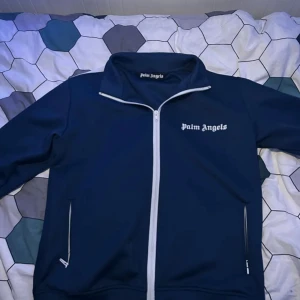 Blå vindjacka från Palm Angels - Snygg marinblå vindjacka från Palm Angels med vit dragkedja och logga på bröstet. Jackan har hög krage och två fickor med dragkedja. Perfekt för dig som gillar streetwear och vill sticka ut med stilren design.