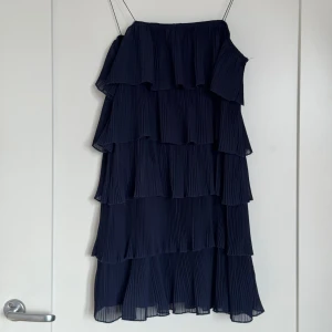 Marinblå plisserad volangklänning - Säljer en marinblå kortklänning med flera lager plisserade volanger. Klänningen är axelbandslös och har en luftig, flowy känsla som ger riktigt snygg rörelse. Perfekt för dig som gillar en cool och trendig look med extra volym. Materialet känns lätt och bekvämt.