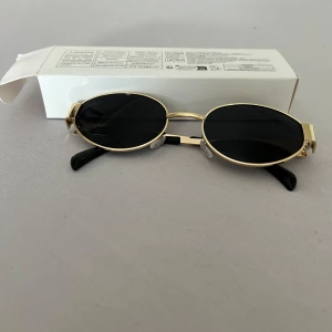 Guldiga ovala solglasögon - Snygga solglasögon med tunna, ovala linser och guldfärgade metallbågar. Svarta glas och svarta detaljer vid skalmarna ger en cool kontrast. Perfekt accessoar för dig som vill sticka ut med en retrovibe och stilren look.