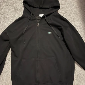 Svart hoodie från Lacoste med dragkedja - Svart hoodie från Lacoste med klassisk krokodillogga på bröstet. Tröjan har huva med snörning, dragkedja framtill och stora fickor. Perfekt för dig som gillar en clean och stilren look.