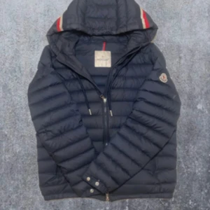 Svart Moncler dunjacka med huva - Svart dunjacka från Moncler med huva och coola röda och vita detaljer på luvan. Jackan har dragkedja framtill, två fickor och klassisk Moncler-logga på ärmen. Puffig design som håller värmen och ger en snygg siluett. Perfekt för kalla dagar.