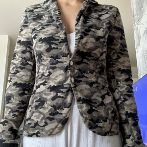 Camouflage kavaj med smal passform - Snygg kavaj i camouflage-mönster med grå, svarta och beige toner. Jackan har smal passform, klassisk krage och knäppning framtill. Perfekt för dig som vill sticka ut med en edgy look. Långärmad och tillverkad i mjukt bomullsmaterial. Ganska liten i storlek - har själv S o den passar