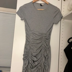 Grå kortklänning från H&M DIVIDED - Snygg grå kortklänning från H&M DIVIDED i ribbad bomull. Klänningen har korta ärmar, rund hals och cool rynkad effekt längs hela framsidan som ger en trendig look. Perfekt för dig som gillar enkel stil med twist.