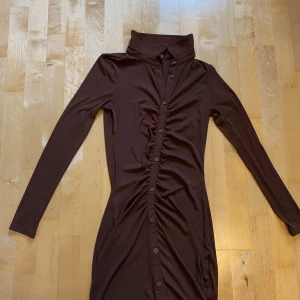 Brun ribbad skjortklänning H&M Divided - Säljer en brun ribbad skjortklänning från H&M Divided i storlek S. Klänningen har lång ärm, markerad krage och knappar hela vägen framtill. Den är figurnära med snygg rynkning längs knappraden och tillverkad i mjuk stretchig polyester.