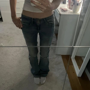 Lågmidjade bootcut jeans med slitningar - Säljer ett par blå lågmidjade jeans med bootcut passform och slitna detaljer vid knäna. Jeansen har klassisk femficksdesign och är gjorda i mjukt denimtyg. Perfekta för dig som gillar en avslappnad och trendig look.