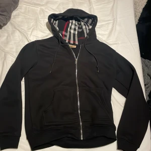 Svart Burberry hoodiejacka med rutor - Svart hoodiejacka från Burberry med dragkedja och klassiskt rutigt foder i huvan. Jackan har snörning i huvan, två fickor fram och mjukt fleecefoder på insidan. Toppskick