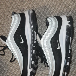Nike Air Max 97 svart/vit sneakers - Nike Air Max 97 sneakers i svart och vitt med ikoniska vågformade linjer och synlig Air-sula. Skorna har svarta snören, vit ovandel med svarta detaljer och en tydlig Swoosh-logga på sidan. Perfekta för dig som gillar en sportig och clean stil.