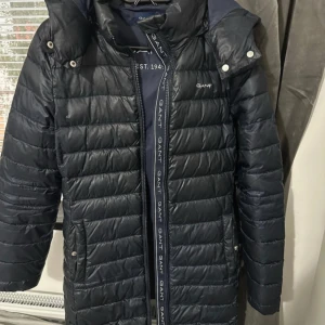 Mörkblå dunjacka från GANT - Snygg mörkblå dunjacka från GANT med huva och dragkedja. Jackan har quiltad design, logga på bröstet och längs dragkedjan samt praktiska fickor med knappar. Perfekt för kyliga dagar och riktigt stilren look. Säljer för 399 men pris kan diskuteras