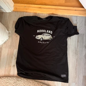 Svart t-shirt Norrland Sortn Company - Svart t-shirt från Sortn Company med tryck av en retrobil och texten 'NORRLAND' på bröstet. Klassisk passform med korta ärmar och rund hals. Skön bomullskvalitet, perfekt för dig som gillar streetwear med cool vintagekänsla.