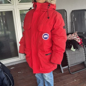 Röd Canada Goose Expedition dunjacka - Säljer en röd Canada Goose Expedition dunjacka med stor huva och pälskant. Jackan har flera stora fickor, patch på bröstet och justerbar dragsko i midjan. Perfekt för kalla vinterdagar och riktigt snygg streetstil. Klassisk logga på bröstet och Expedition-märke på ryggen. Äkta. Xs på den men passar upp till M ( för stor på mig som är 1.57 lång och xs/s)