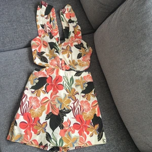 Blommiga shorts med hög midja - Snygga högmidjade shorts med tropiskt blommönster i färger som röd, gul, grön och svart på en ljus bas. Shortsen har en skön passform och breda ben, perfekta för varma sommardagar. Materialet känns som bomull och ger en luftig känsla.