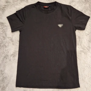 Svart t-shirt från Prada med logga - Svart t-shirt från Prada med klassisk rund hals och korta ärmar. På bröstet sitter en ikonisk metalltriangel med Prada Milano-loggan. T-shirten har en clean och stilren look, perfekt för dig som gillar designerplagg med diskreta detaljer. Materialet känns mjukt och är troligtvis bomull. Priset kan diskuteras. Den passar Xs, s och även M