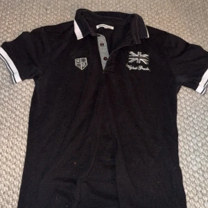 Svart pikétröja med broderi - Svart pikétröja från Highest Grade med broderad brittisk flagga och text på bröstet. Har vita detaljer på krage och ärmslut samt knappslå med kontrastfärg. Perfekt för dig som gillar klassisk stil med en twist.