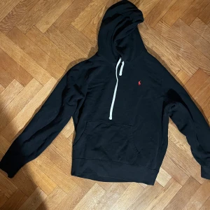 Mörkblå hoodie från Ralph Lauren - Mörkblå hoodie från Ralph Lauren med vit snörning i huvan och klassisk röd logga på bröstet. Tröjan har känguruficka framtill och långa ärmar. .
