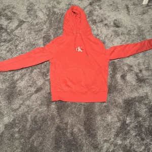 Röd hoodie från Calvin Klein Jeans - Snygg röd hoodie från Calvin Klein Jeans med klassisk CK-logga på bröstet. Tröjan har huva med snörning, magficka och långa ärmar. Perfekt för dig som gillar streetstyle och vill ha en bekväm och trendig look.