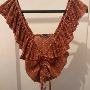 Prickig volangblus från Zara - Supersöt brun blus från Zara med vita prickar, djup v-ringning och breda volanger längs axlarna. Toppen har snörning framtill för en snygg rynkad effekt och är ärmlös. Perfekt för dig som gillar unika detaljer och trendig stil.