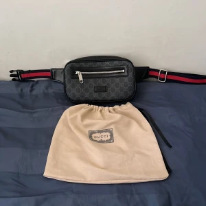 Svart midjeväska - Säljer en snygg svart midjeväska med klassiskt GG-monogram och silverfärgad dragkedja. Väskan har justerbart band i rött och marinblått, samt detaljer i skinn. Perfekt för att bära det viktigaste och ger en lyxig touch till din outfit. Påsen ingår inte!