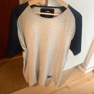 Grå t-shirt med marinblå ärmar Core - Snygg t-shirt från Jack & Jones Core med ljusgrå framsida och baksida samt kontrasterande marinblå raglanärmar. Klassisk rund halsringning och normal passform. Perfekt för en avslappnad stil.
