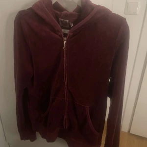 Vinröd velour hoodie från Juicy Couture - Juicy Couture hoodie i vinröd velour med guldfärgad dragkedja och huva. Ikonisk logga med strass på ryggen och fickor framtill. Perfekt för dig som gillar Y2K-vibe och comfy stil. Snygg att matcha med jeans eller mjukisbyxor.