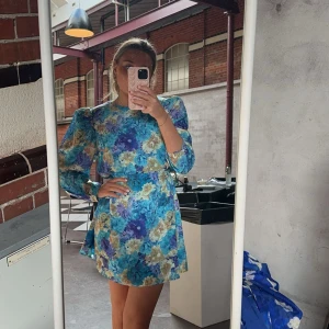 Blommig blå kortklänning från Adoore - Supersöt kortklänning från Adoore med blommigt mönster i blå, lila och beige toner. Klänningen har puffiga långärmar, rund hals och markerad midja med knytband. Materialet känns lätt och flowy, perfekt för att sticka ut med färg och mönster.