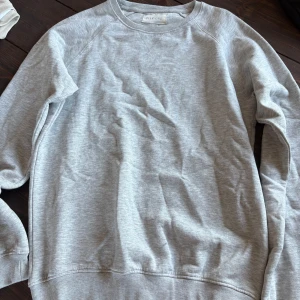 Ljusgrå sweatshirt från Pier One - En stilren ljusgrå sweatshirt från Pier One med klassisk rund halsringning och ribbade muddar. Tröjan är i mjuk bomullsmix och har en avslappnad passform, perfekt för en chill och enkel look.