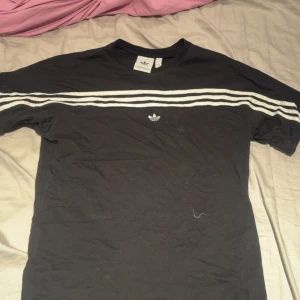 Svart Adidas t-shirt med vita ränder - Svart t-shirt från Adidas med klassiska tre vita ränder över bröstet och broderad Adidas-logga framtill. Rund hals och korta ärmar. Perfekt för dig som gillar sportig och clean stil.