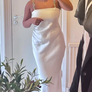 Vit långklänning med kedjeband - Säljer en elegant vit långklänning i satinliknande material med raka linjer och smala axelband i färgglad kedjedesign. Klänningen har en stilren, figurnära passform och är perfekt för dig som vill sticka ut med en unik detalj på axelbanden.