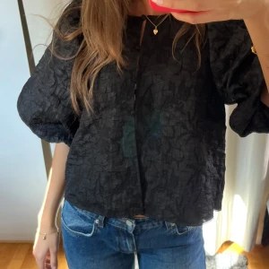 Svart blus med puffärmar - Säljer en svart blus med coolt, krinklat tyg och breda puffärmar. Blusen har en lös passform och en liten öppning med knyt fram vid halsen.  Aldrig använd!