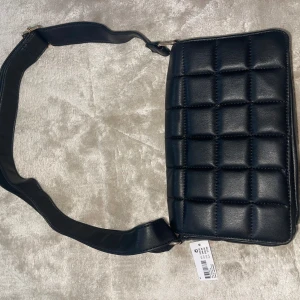 Svart quiltad axelväska i skinnimitation - Stilren svart axelväska med quiltad design och bred axelrem. Väskan har en rektangulär form och är tillverkad i skinnimitation. Perfekt för att lyfta vilken outfit som helst med en modern touch.