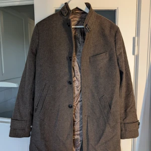 Grå ullrock från Riley Stanton - Snygg grå rock från Riley Stanton med klassisk rak passform och hög krage med spänne. Jackan har knäppning framtill, flera fickor och är helfodrad med lyxig känsla. Perfekt för dig som vill ha en stilren och tidlös look under kyliga dagar.