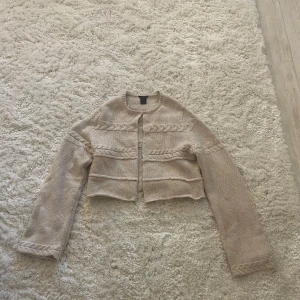 Beige stickad kofta från Lindex - Superfin beige stickad kofta från Lindex i croppad modell. Koftan har flätade detaljer längs ärmar och över bröstet, rund halsringning och långa ärmar. Perfekt nu till hösten 🍂