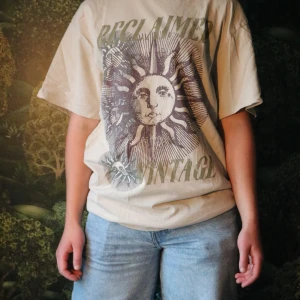 Beige Reclaimed Vintage t-shirt - Säljer en oversized beige t-shirt från Reclaimed Vintage med coolt soltryck och text på framsidan. T-shirten har korta ärmar, rund hals och är gjord i mjuk bomull. Perfekt för en avslappnad och trendig look. Lappen är bortklippt
