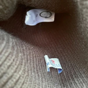 Mörkbrun ribbstickad Moncler mössa - Snygg mörkbrun ribbstickad mössa från Moncler med klassisk uppvikt kant och broderad logotyp framtill. Perfekt för kalla dagar och ger en clean, exklusiv look. Tillverkad i mjukt ullmaterial som håller dig varm hela vintern. Pris diskuteras