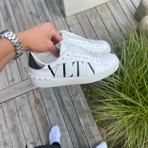 Valentino Garavani VLTN vita sneakers - Snygga vita sneakers från Valentino Garavani med stor svart VLTN-logga på sidan. Skorna har silvriga detaljer på hälen, nitar runt sulan och är gjorda i skinn. Klassisk rund tå och snörning framtill. Perfekta för dig som gillar exklusiv streetstyle. Påse och exktra skosnören fås med