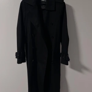 Svart trenchcoat från Mango - Stilren svart trenchcoat från MNG Selection med klassisk dubbelknäppning, bälte i midjan och spännen vid ärmsluten. Jackan är vattenavvisande och har en snygg, lång siluett som ger en clean och tidlös look.