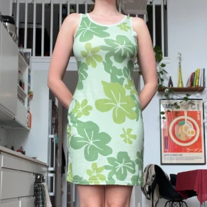 Grön blommig kortklänning - Säljer en söt ärmlös kortklänning med grönt blommigt mönster på ljus bakgrund. Klänningen har rund hals med vit spetskant.  