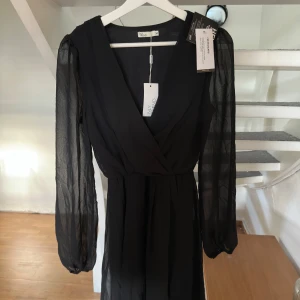 Svart omlottklänning från NLY Trend - Snygg svart omlottklänning från NLY Trend i storlek 34. Klänningen har transparenta långärmade ärmar, markerad midja och v-ringad hals. Materialet är lätt och ger ett elegant fall. Perfekt för dig som vill ha en stilren look med lite edge.
