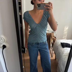 Blå v-ringad topp från Free People - Säljer en blå topp från Free People med v-ringning och rynkade detaljer längs mitten och axlarna. Toppen har korta ärmar och dekorativ kant runt halsen. Materialet är mjukt och lätt, perfekt för varma dagar. Supersnygg att styla med jeans eller kjol!